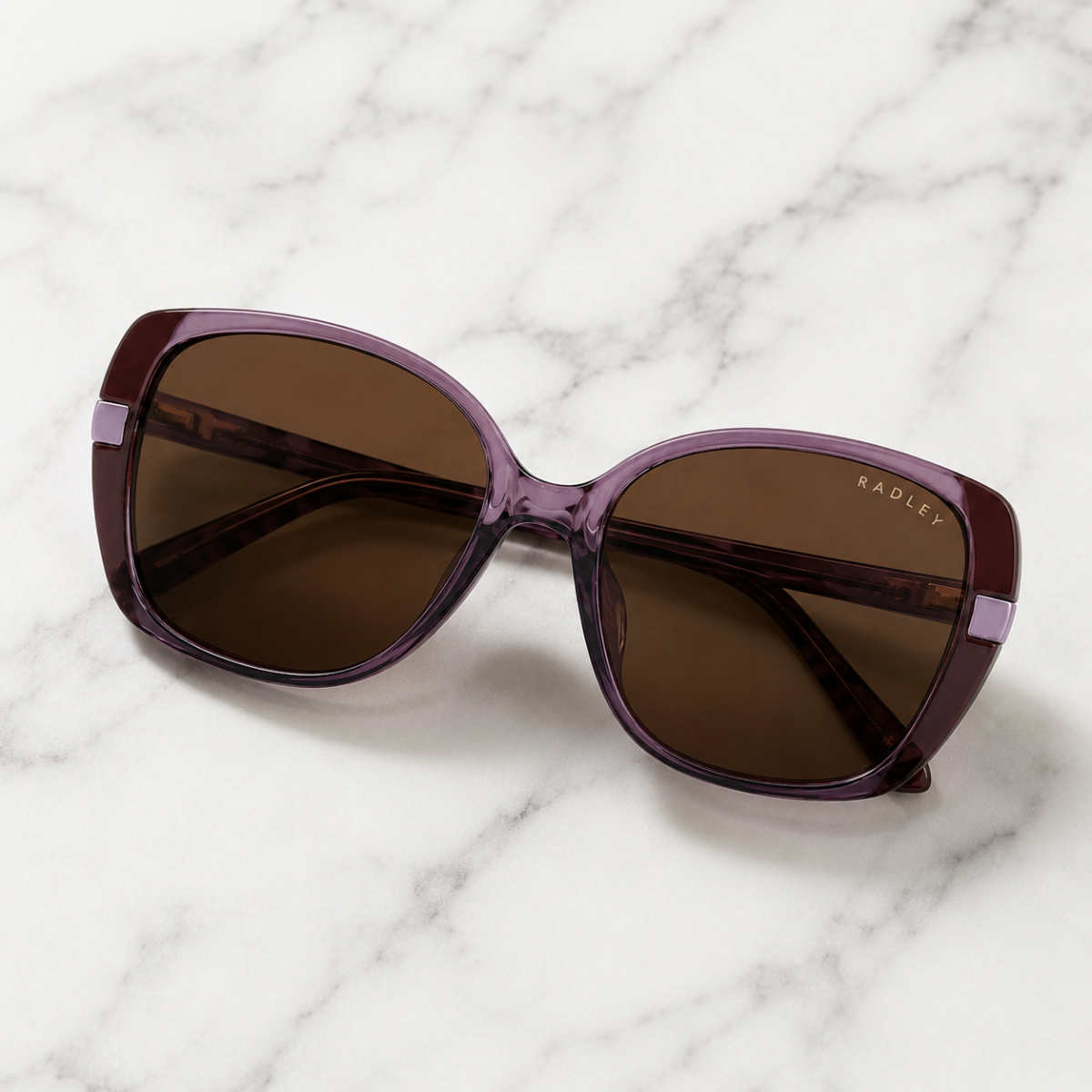 Radley sunglasses with a translucent purple acetate frame and dark brown lenses, resting on a white marble surface.